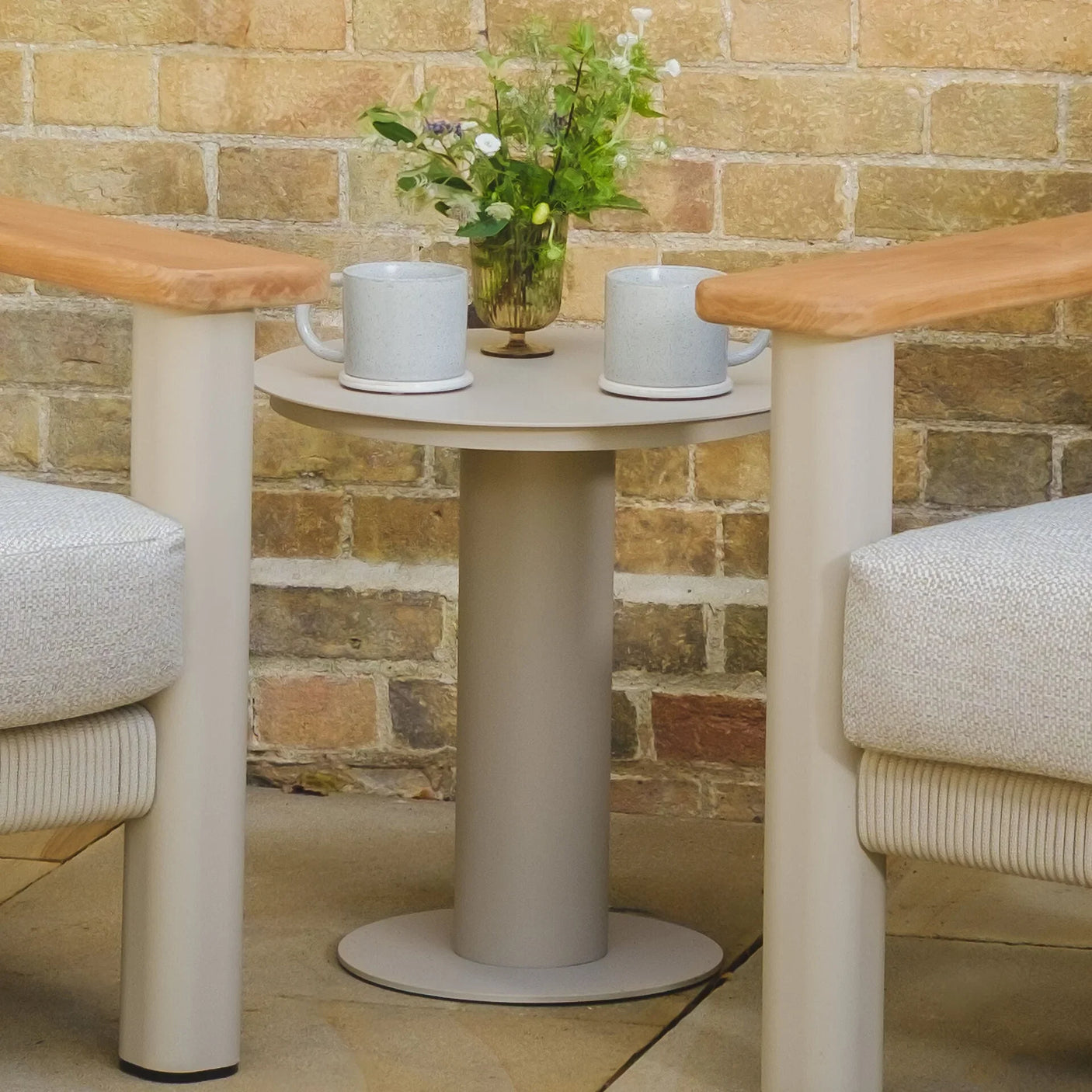 Round outdoor side table with grey mugs and floral vase between two patio chairs