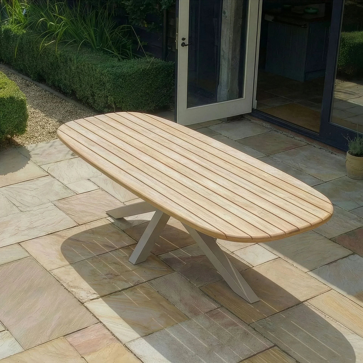 Wooden outdoor dining table with slatted top on stone patio near garden and open door
