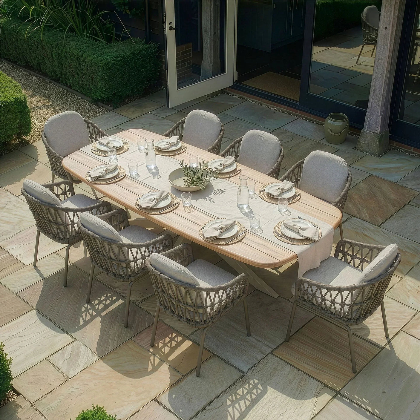 Outdoor dining set with light wood table, eight cushioned rattan chairs, and neutral tableware