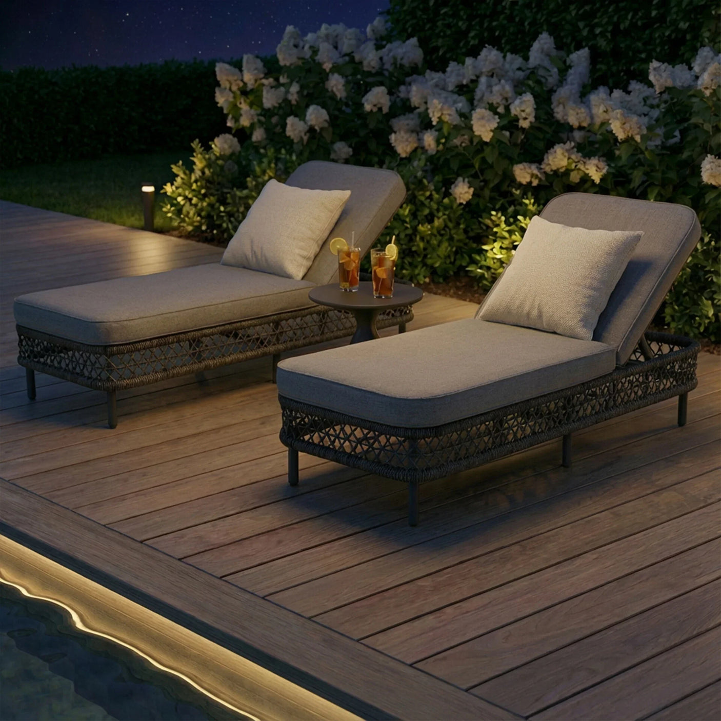 Rattan sun loungers with grey cushions and side table on deck by pool at night