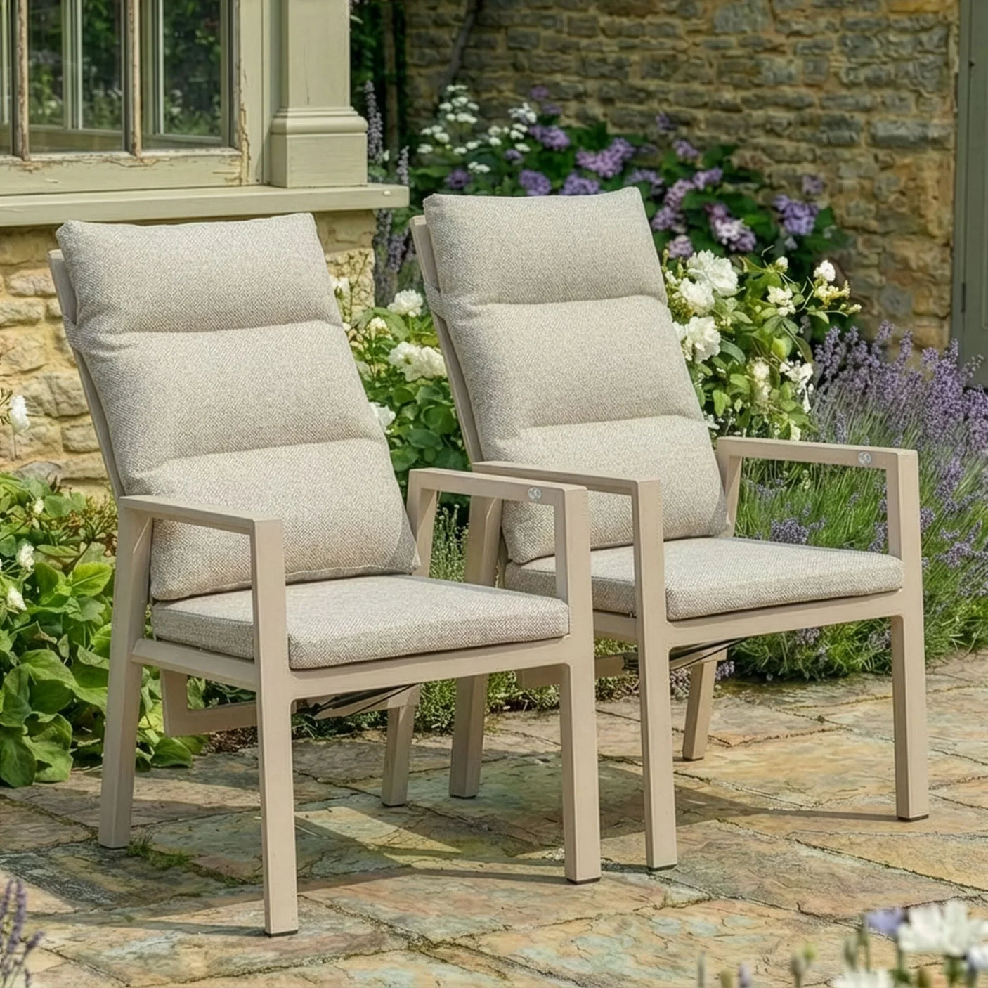 Pair of beige cushioned garden chairs with armrests on stone patio surrounded by flowers