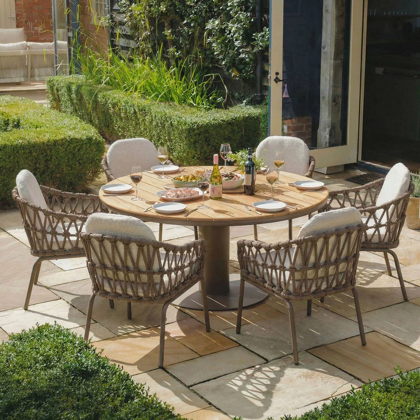 Round wooden outdoor dining table set with six rattan chairs and neutral cushions on patio