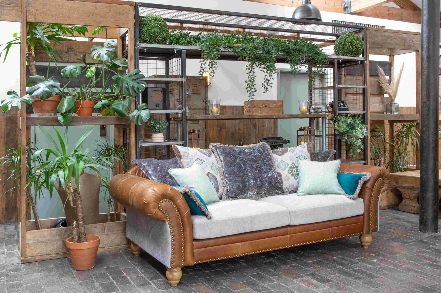 Brown leather and fabric sofa with assorted cushions, surrounded by indoor plants and wooden shelves
