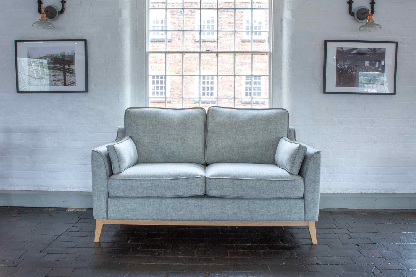 Light grey fabric sofa with wooden legs against white brick wall and large window