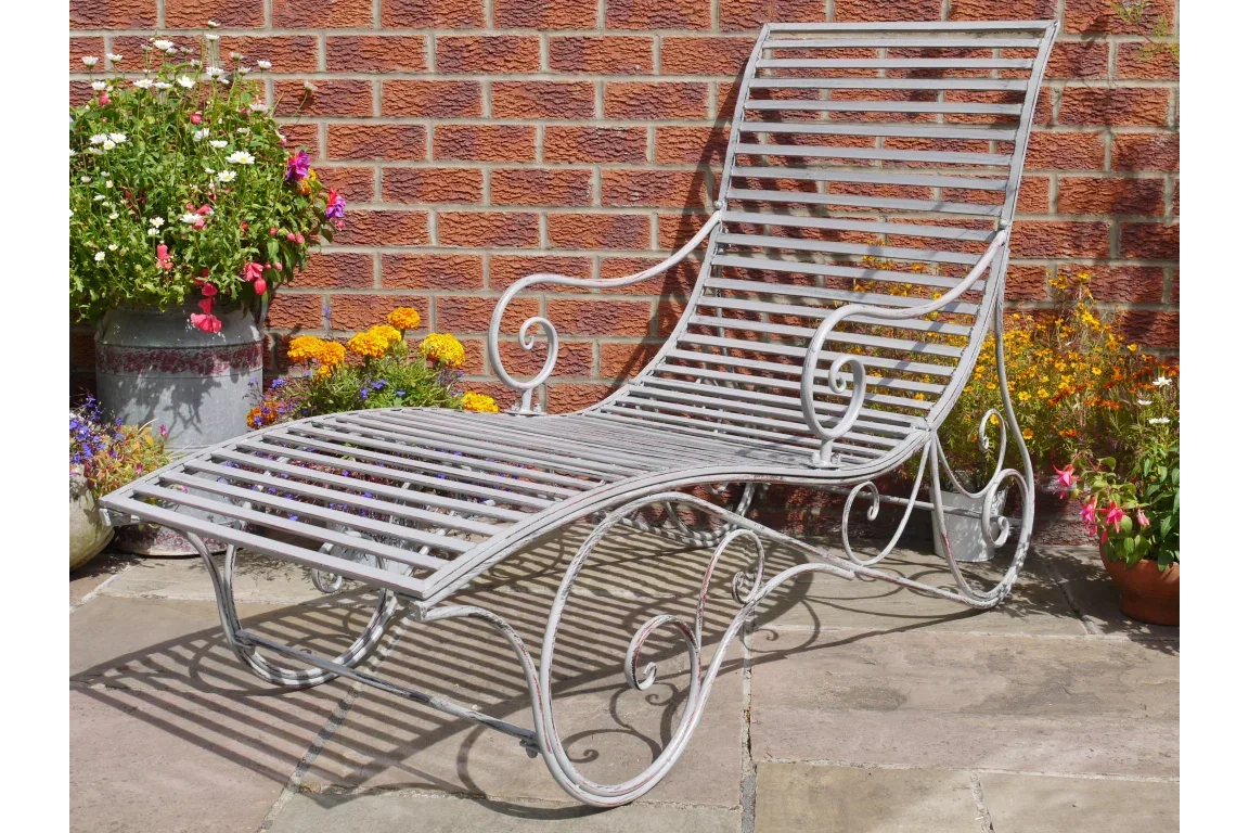 metal garden lounger with slatted seat and scroll armrests on patio by brick wall and flowers