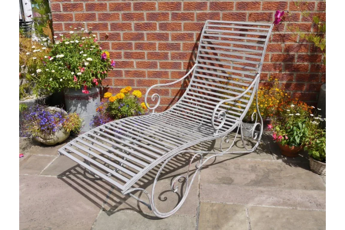 Grey metal garden lounger with slatted design and scroll arms on patio near brick wall