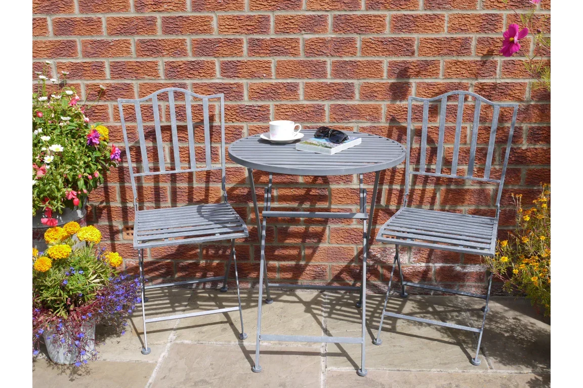 Metal bistro set with round table and two chairs on patio by brick wall, flowers nearby