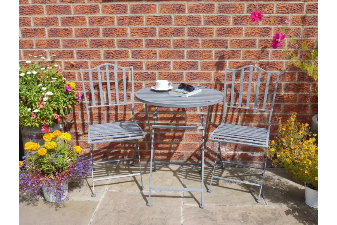 Metal bistro table and two chairs set on patio with flowers and brick wall background
