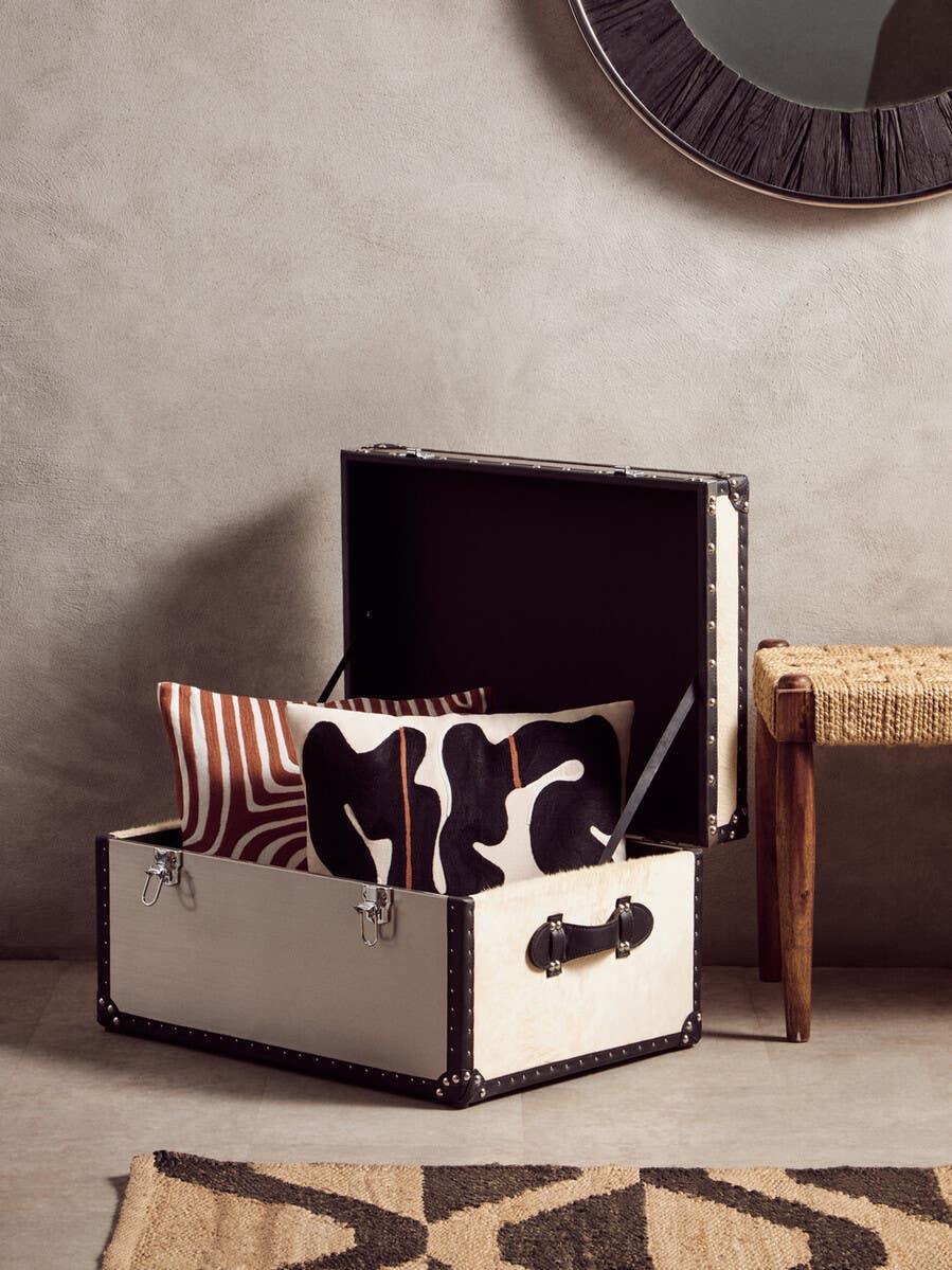 White storage trunk with black trim, open, holding patterned cushions beside rustic bench