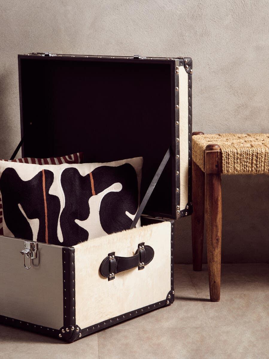 Storage trunk with black trim and handle, patterned cushion inside, next to woven bench