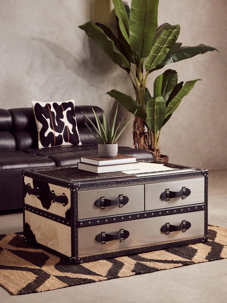 Metal trunk coffee table with drawers, on geometric rug, beside black leather sofa and plants