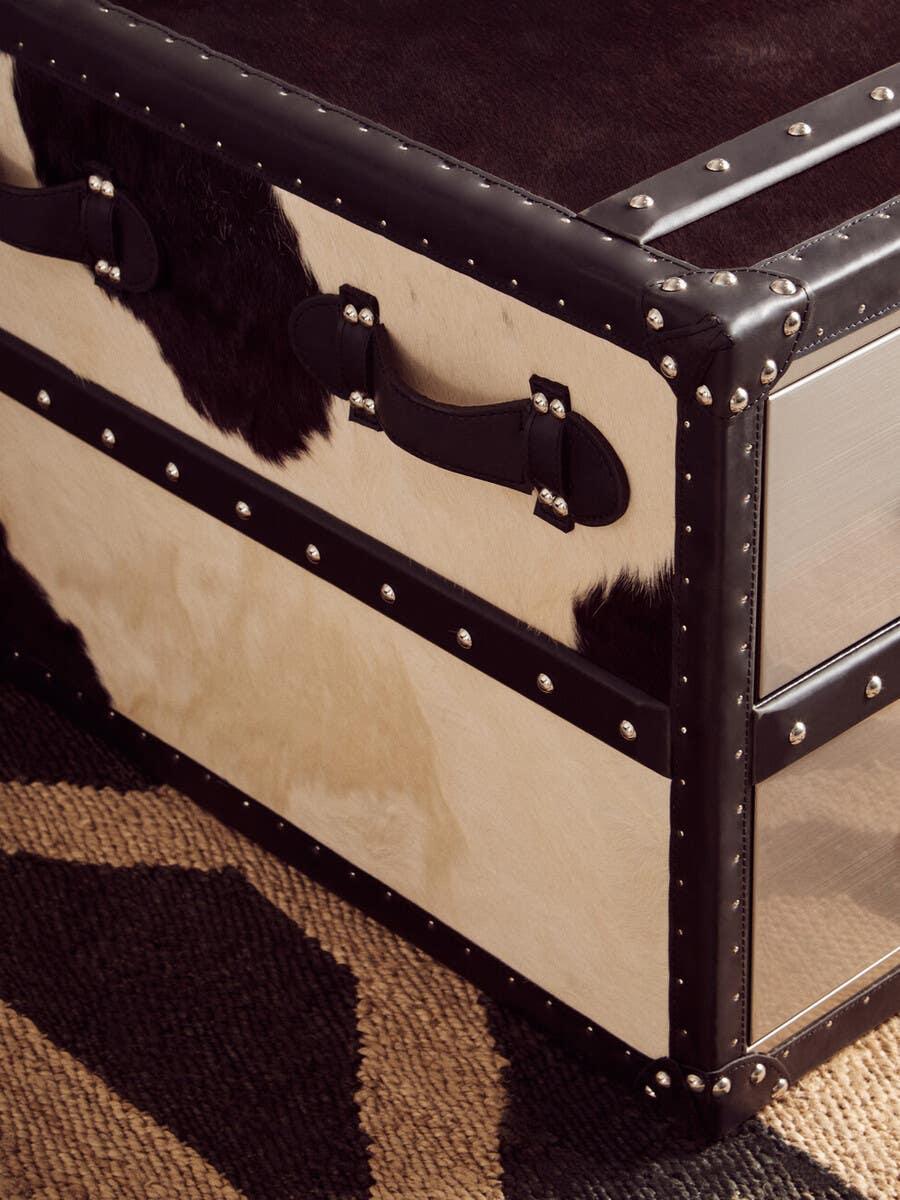 Cowhide storage trunk with black leather handles and metal stud detailing on patterned rug