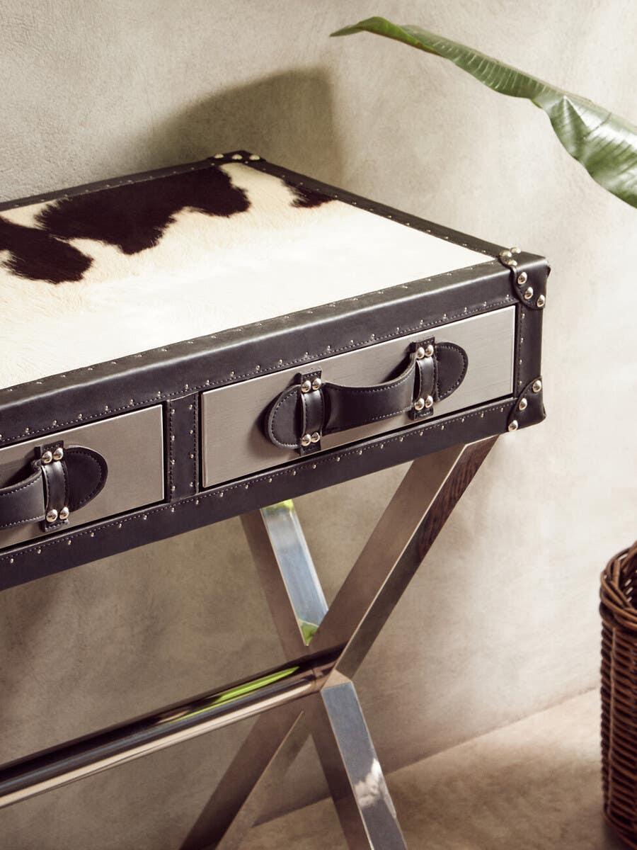 Cowhide console table with two front drawers and chrome crossed legs beside potted plant