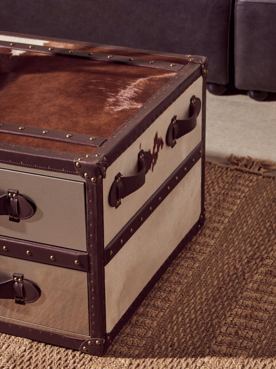 Cowhide storage trunk with leather trim and handles on woven rug beside dark sofa