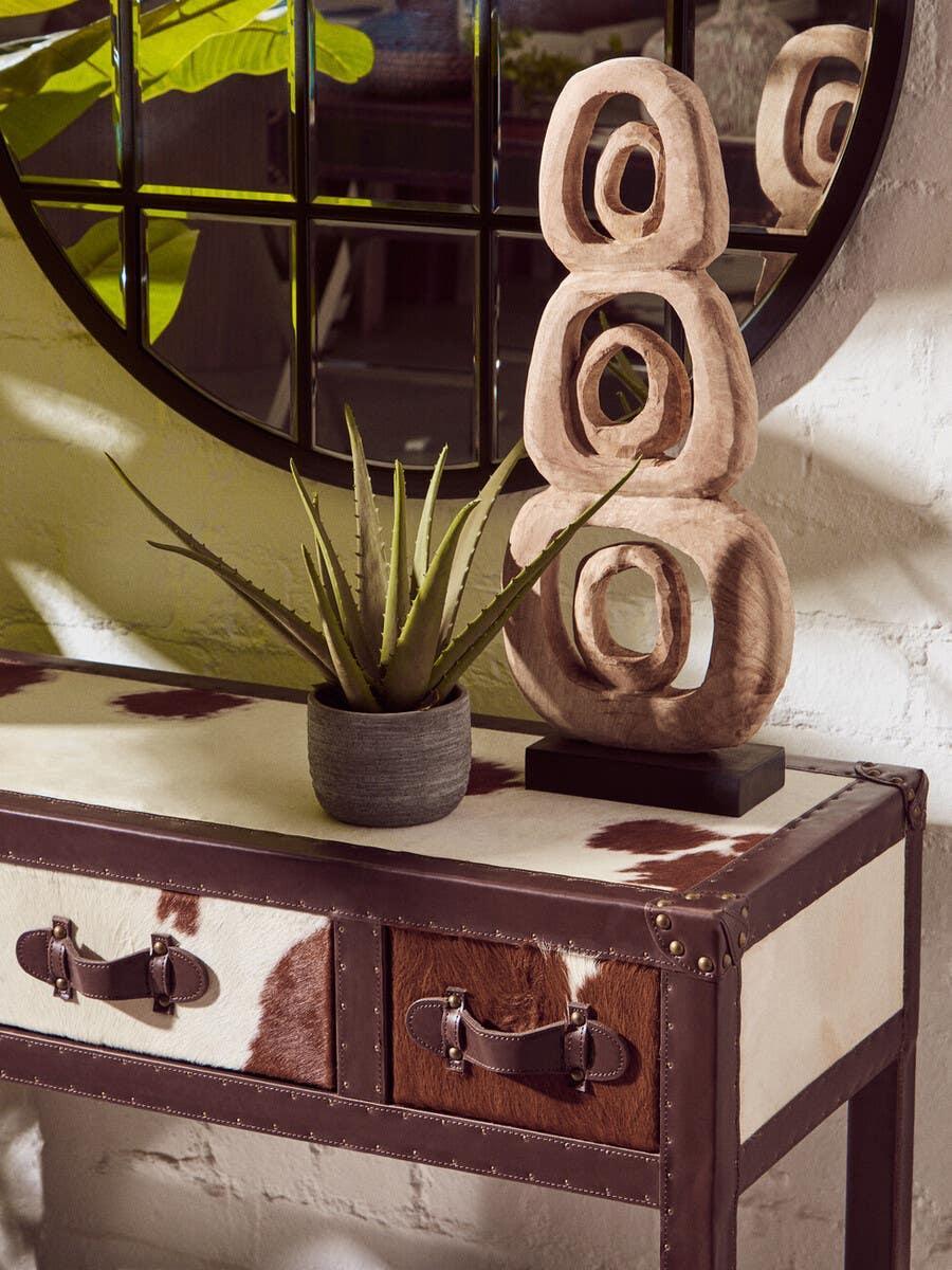 Cowhide console table with leather trim, decorative sculpture and plant, mirror in background