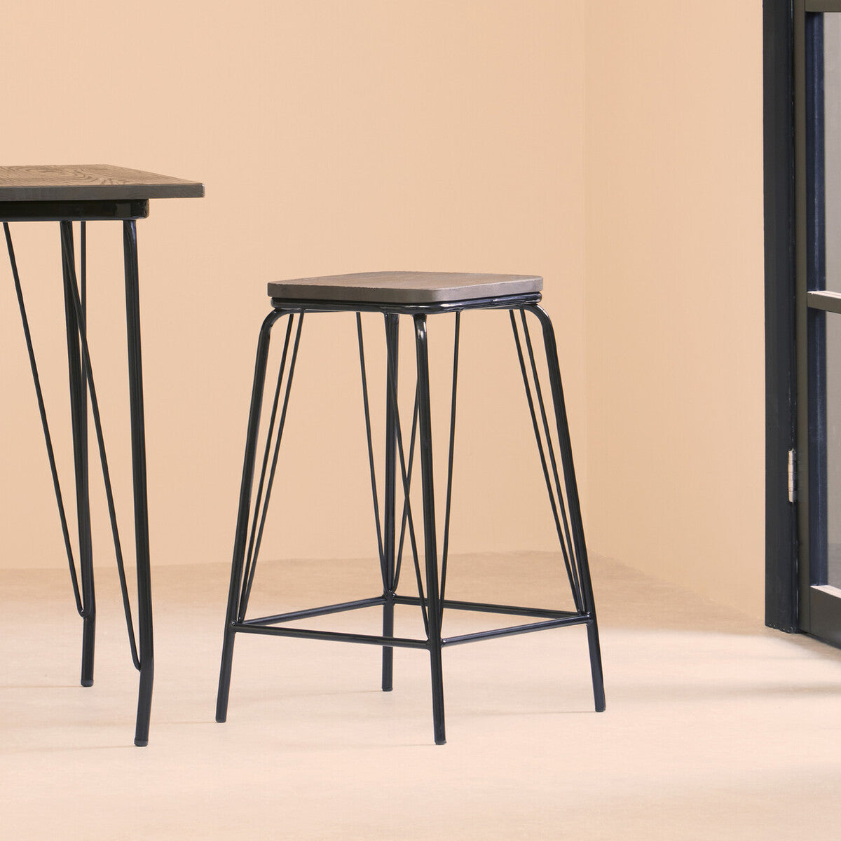 A black metal stool with a simple design featuring a square elm wood seat, supported by slender, angled legs. The stool is positioned next to a matching table against a light-colored wall, creating a modern and minimalist aesthetic