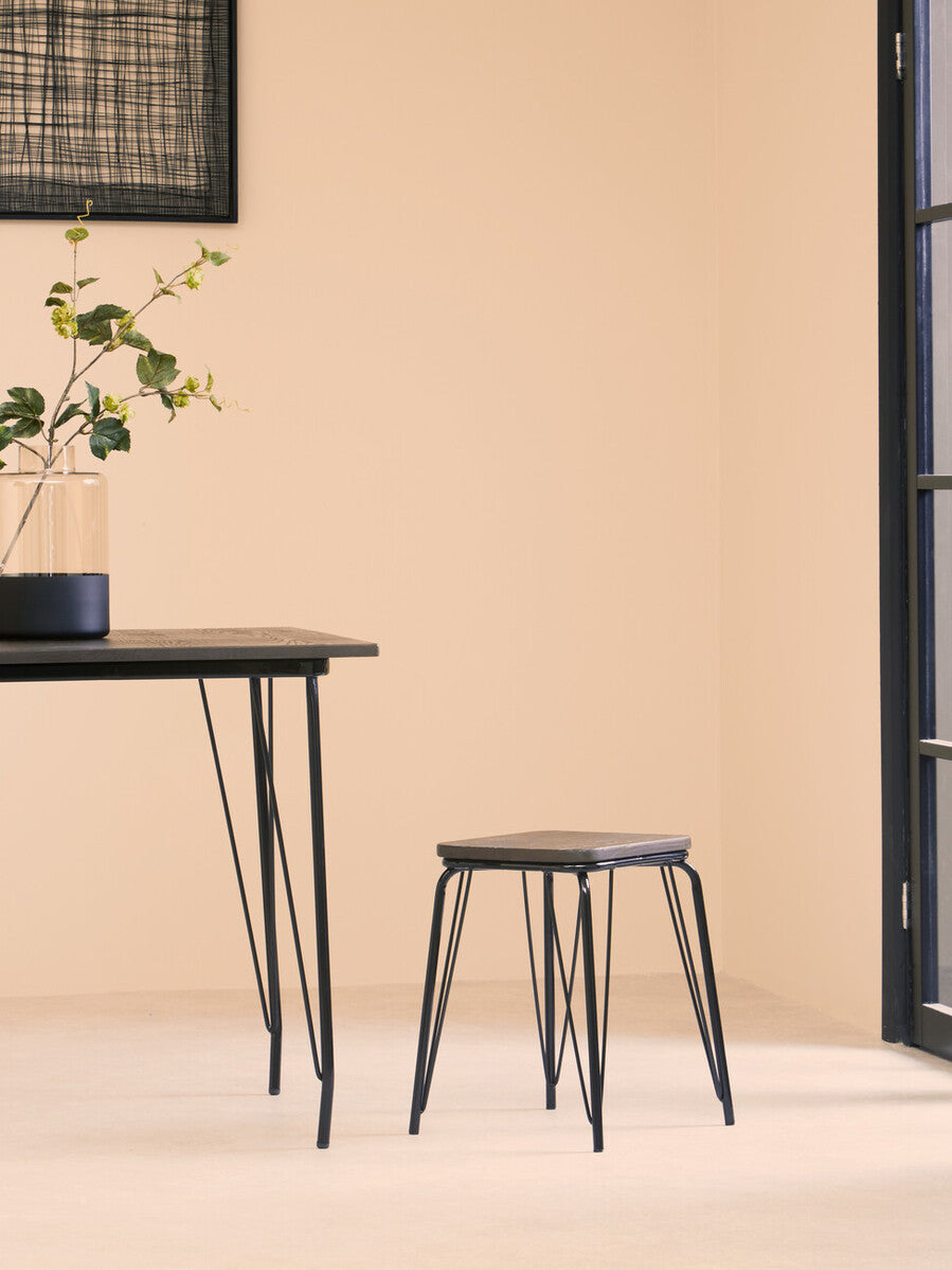 A small black metal stool with a wooden elm seat is positioned next to a sleek black table in a softly lit room with beige walls. A potted plant sits on the table, adding a touch of greenery to the minimalist decor. Natural light streams in through a large window, enhancing the warm atmosphere