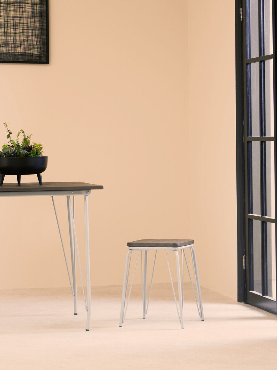 A small wood stool featuring a grey metal frame and a natural elm seat, positioned next to a sleek table in a minimalist room with light-colored walls and a large window. A small potted plant sits on the table, adding a touch of greenery to the scene