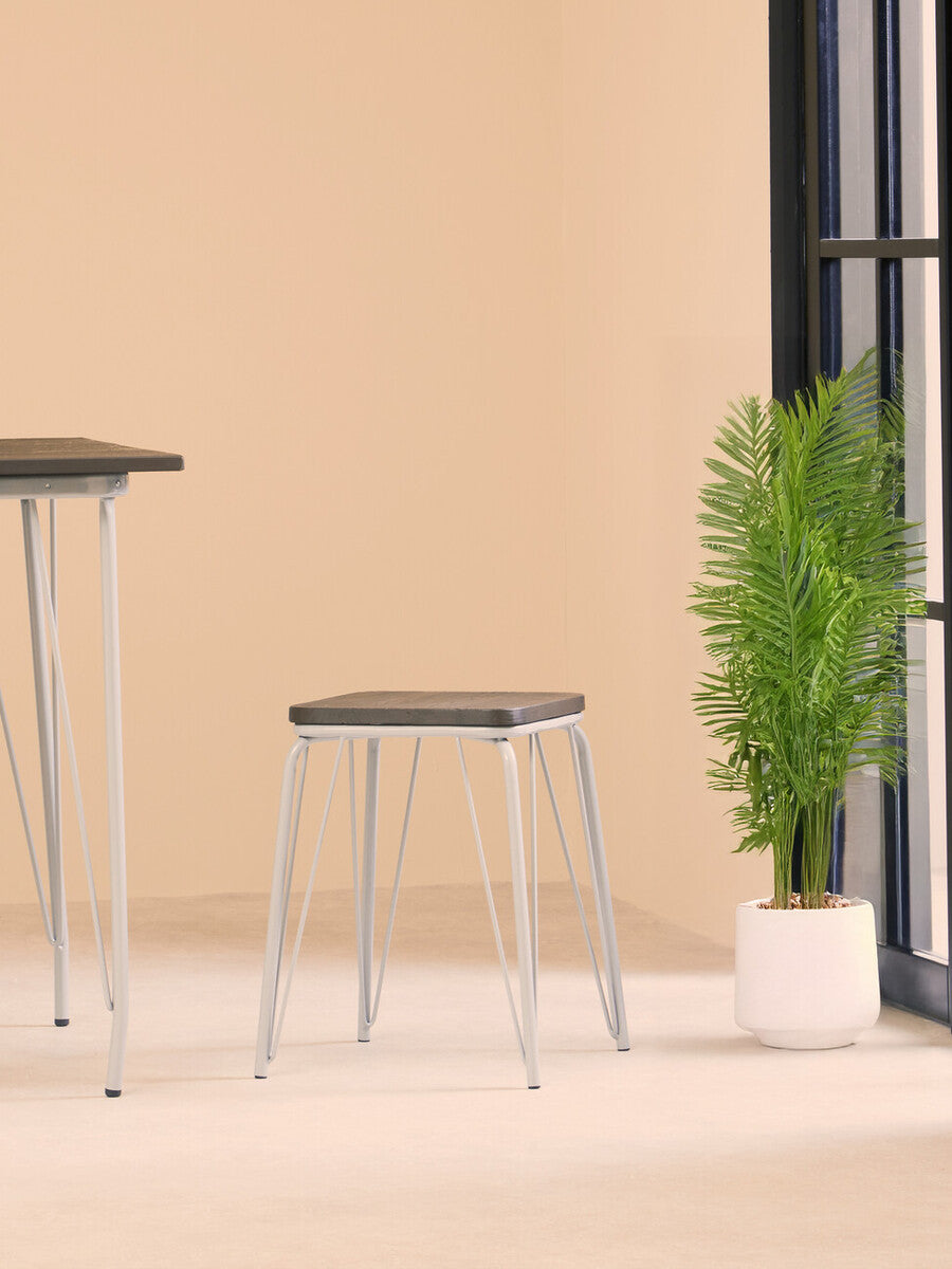 Small wood stool featuring a grey metal frame and an elm wood seat, positioned on a light-colored floor with a potted plant in the background against a beige wall