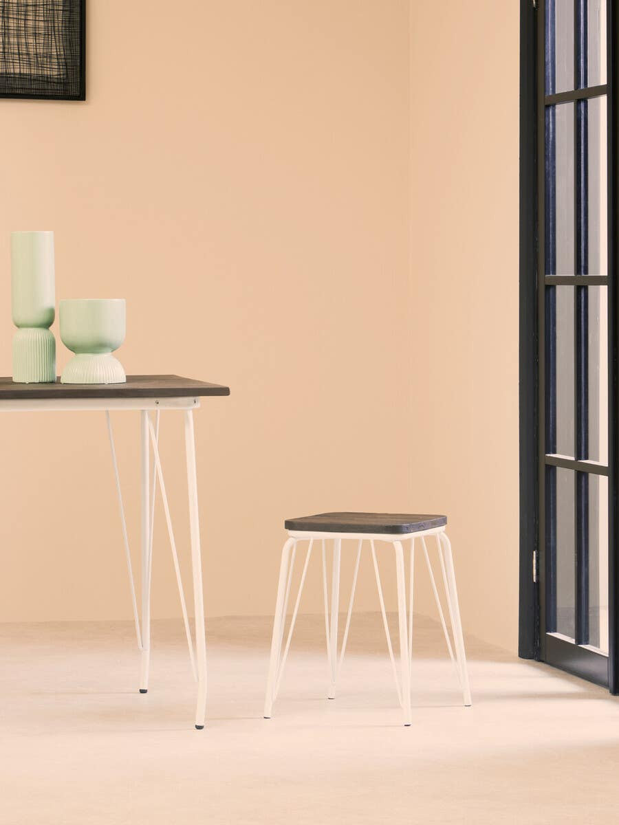 Small wood stool with a dark elm seat and white metal legs, positioned next to a light-colored table. The background features a soft beige wall and a large window with black frames, allowing natural light to illuminate the scene. A few decorative items in soft green tones are placed on the table