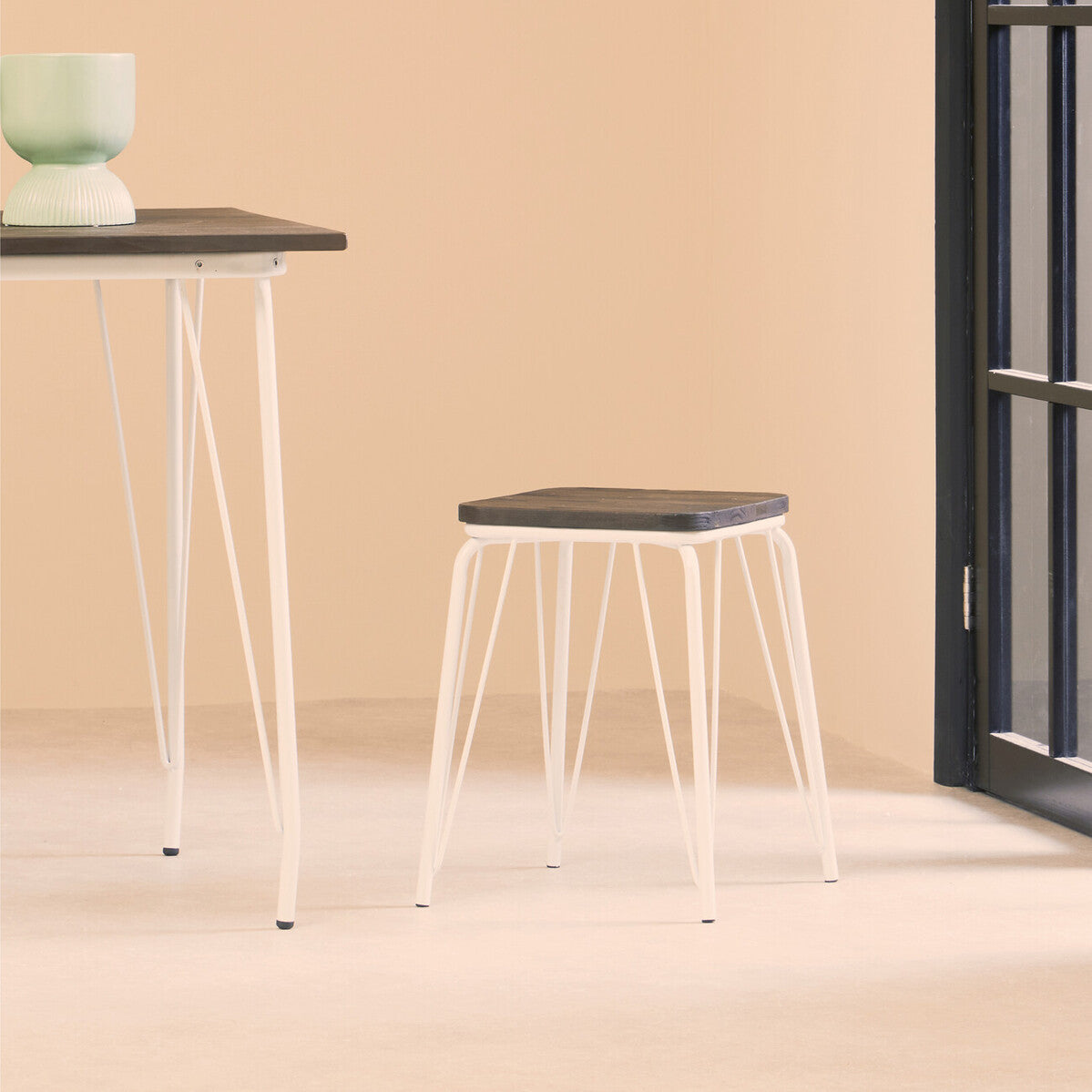 Small wood stool with a dark elm seat and white metal legs, positioned next to a matching table in a softly lit room with a light-colored wall and a large window