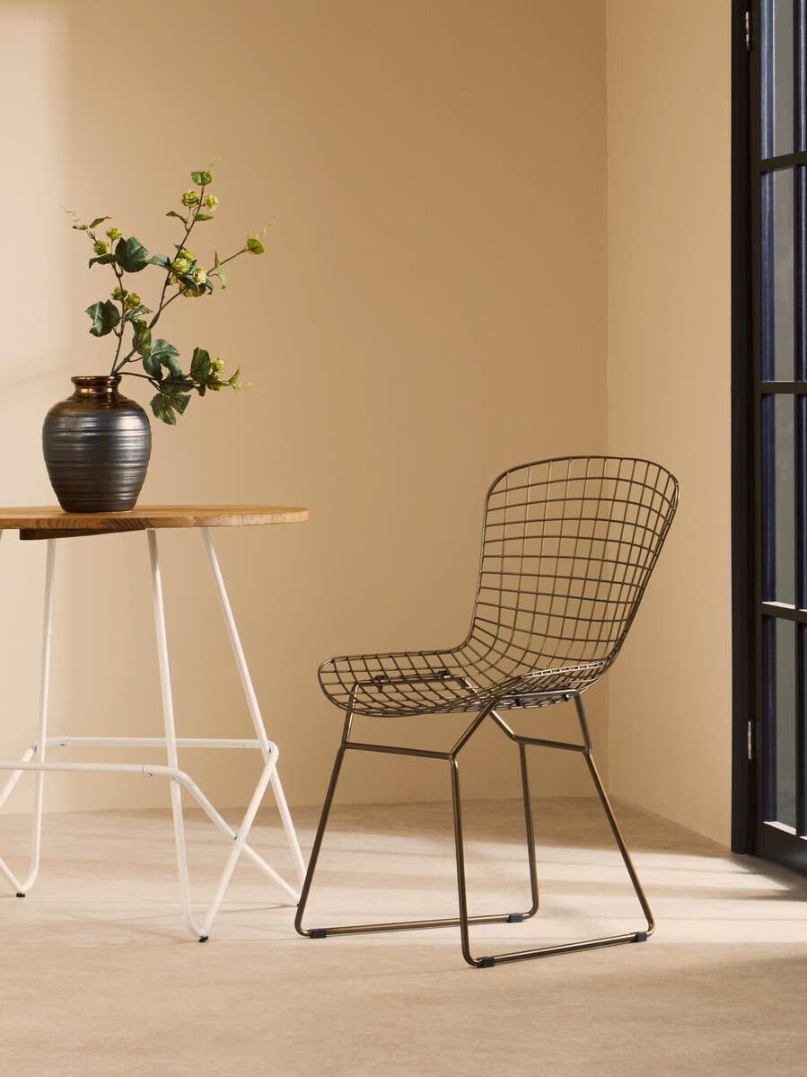 Bronze metal grid frame wire chair positioned next to a small wooden table with a vase of flowers, set against a light beige wall and large window