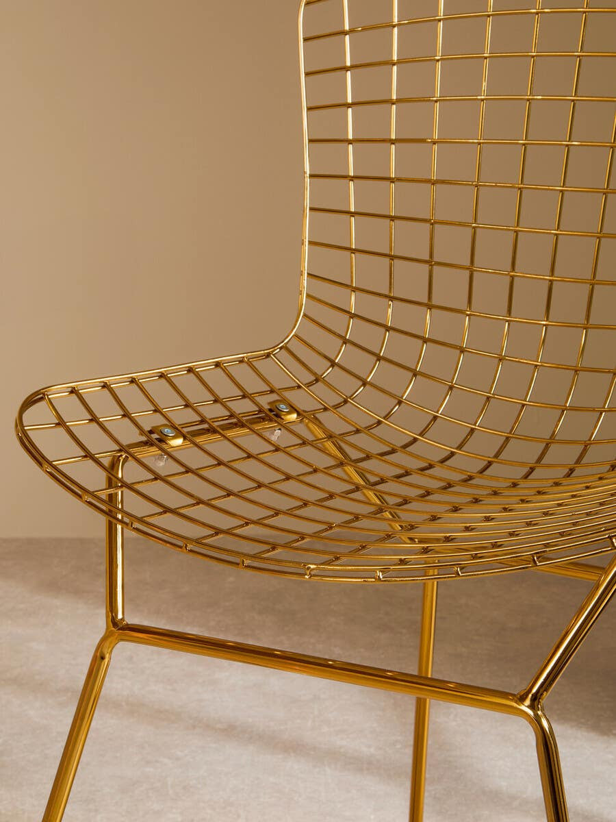 A close-up view of a gold metal grid frame wire chair, showcasing its sleek, modern design with a curved seat and backrest made of thin metal wires, supported by a minimalist base. The background features a soft, neutral wall color that complements the chair's metallic finish