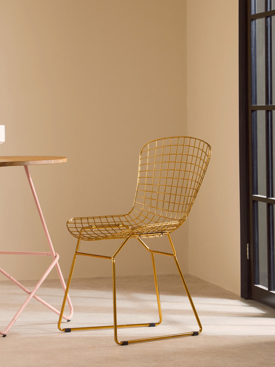 A gold metal grid frame wire chair with a minimalist design, positioned next to a light-colored table in a softly lit room with beige walls. The chair features a curved backrest and a simple, elegant silhouette, complementing the modern decor