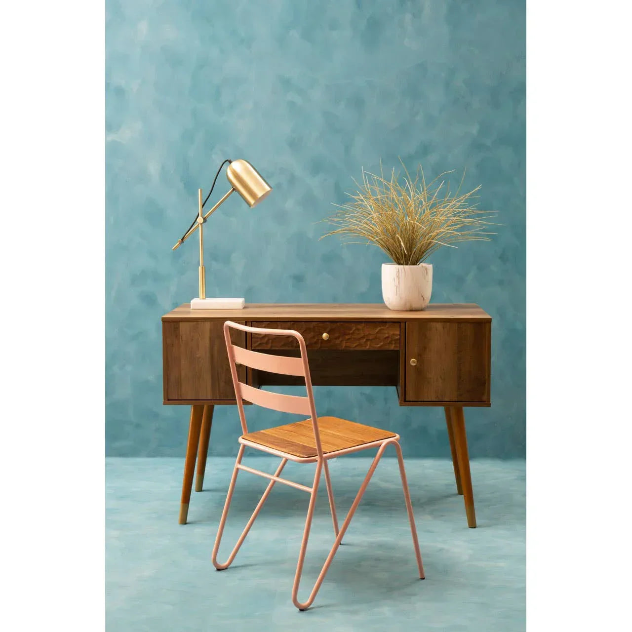 Mid-century wood desk with gold lamp, potted plant, and pink metal chair against blue wall