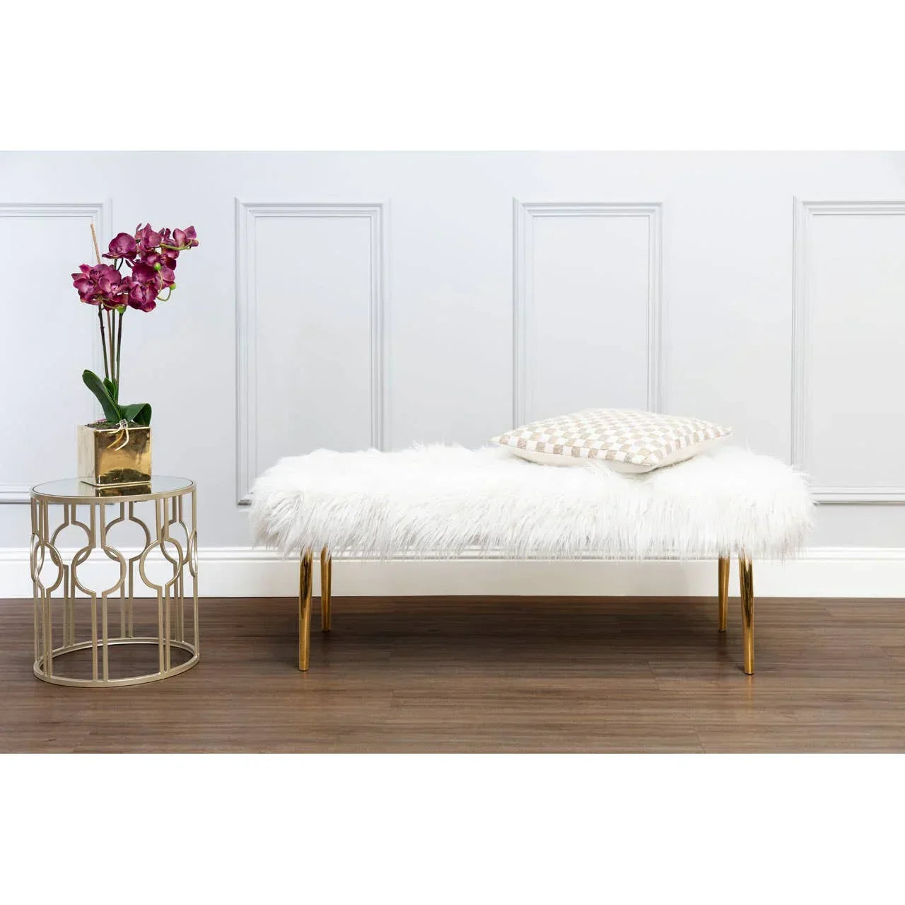 White faux fur bench with gold legs and cushion beside gold side table with orchid