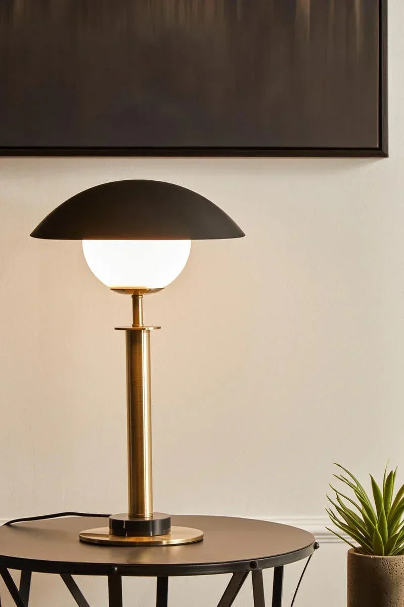 Modern brass table lamp with matte black dome shade on round side table next to plant