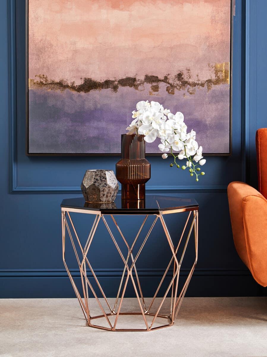 Rose gold metal side table with glass top, geometric base, and decorative vases in modern room