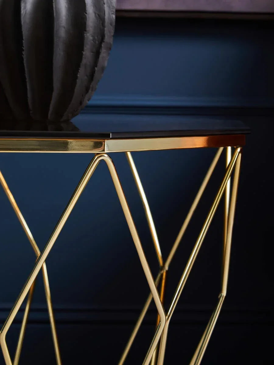 Gold metal side table with geometric legs and black glass top against dark blue wall