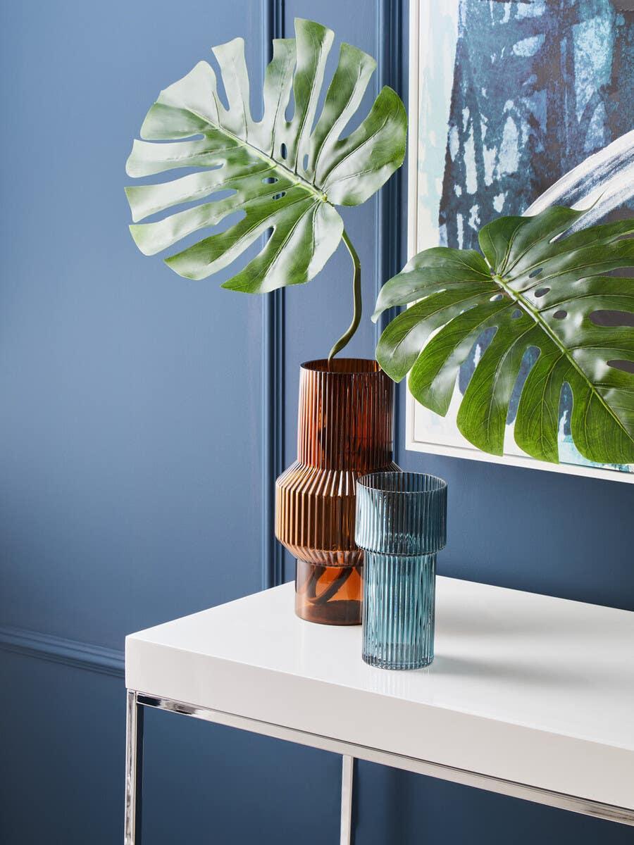 Glass vases with artificial monstera leaves on a white console table against a blue wall