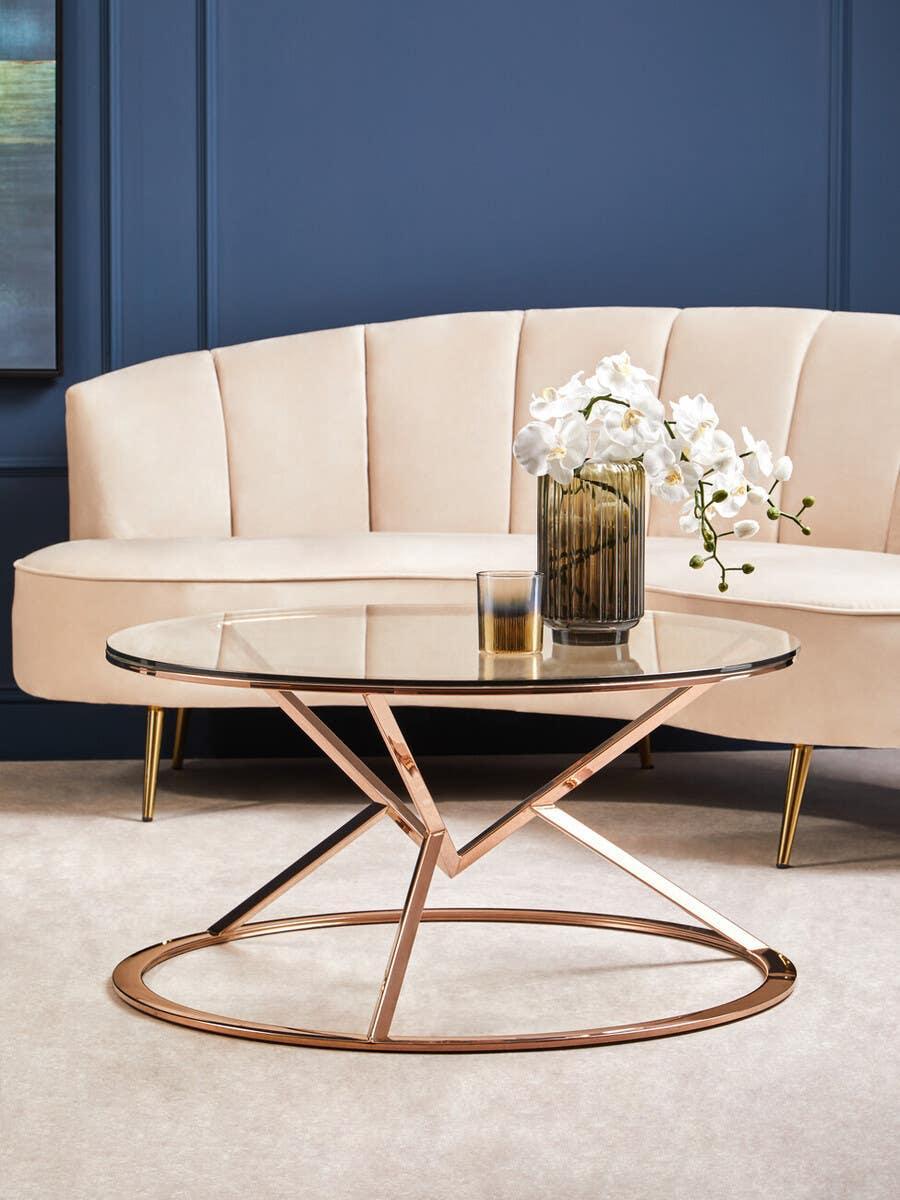 Glass coffee table with rose gold metal frame in front of cream velvet sofa and blue wall