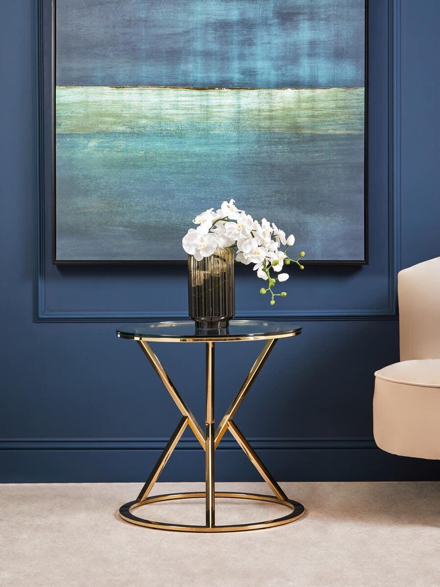 Round gold side table with glass top and vase on blue wall background