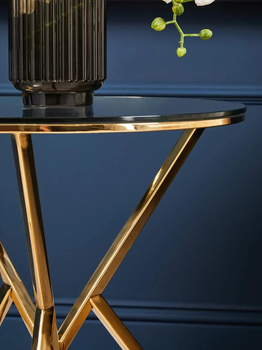 Round glass side table with gold metal legs, black vase on top against blue wall