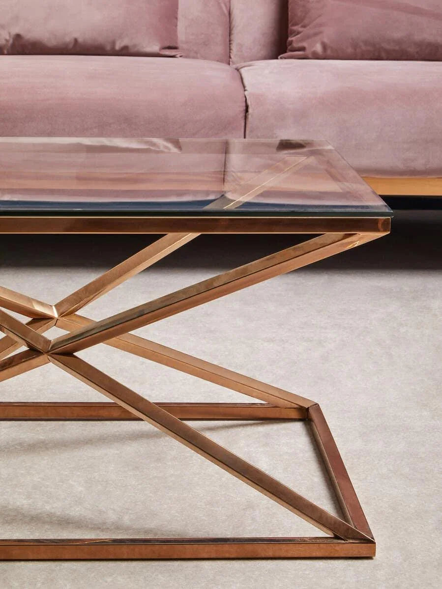 Glass top coffee table with geometric gold metal base in front of pink velvet sofa