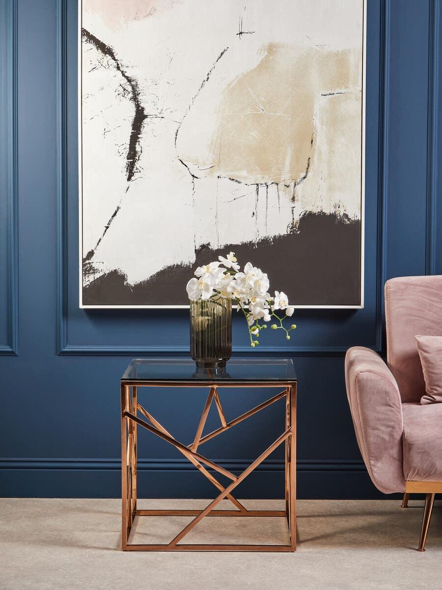 Modern gold metal side table with glass top beside blush velvet armchair and abstract art