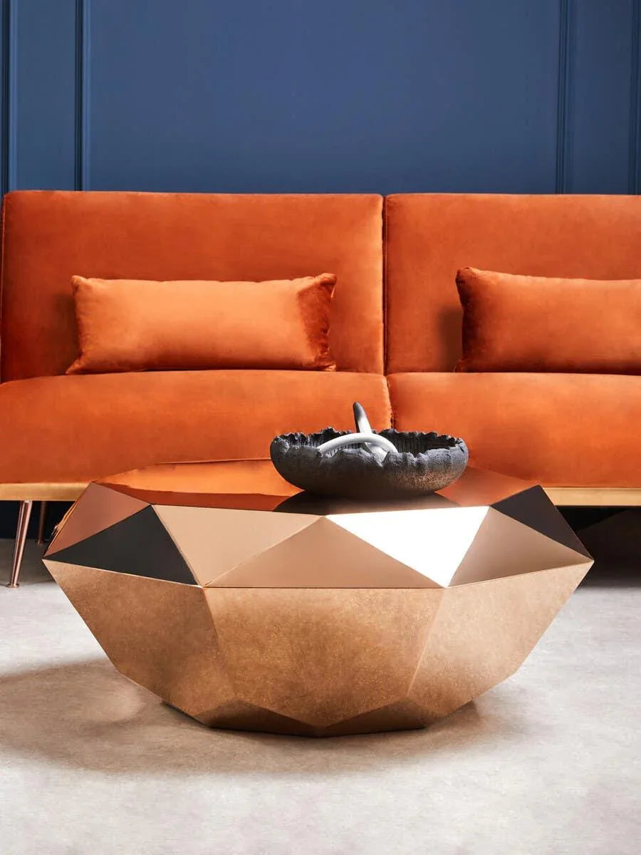 Geometric gold coffee table with decorative bowl in front of orange velvet sofa and blue wall
