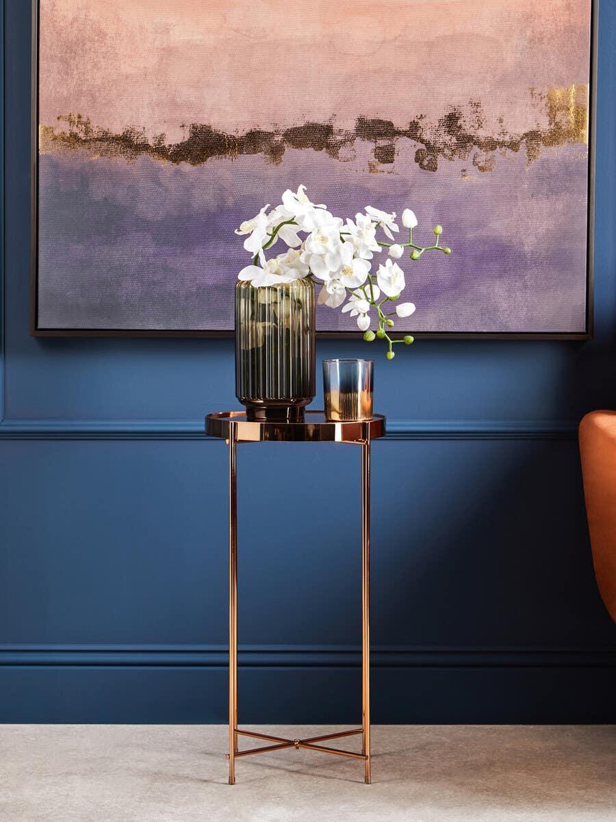 Round gold side table with glass vase and white orchid flowers against blue wall