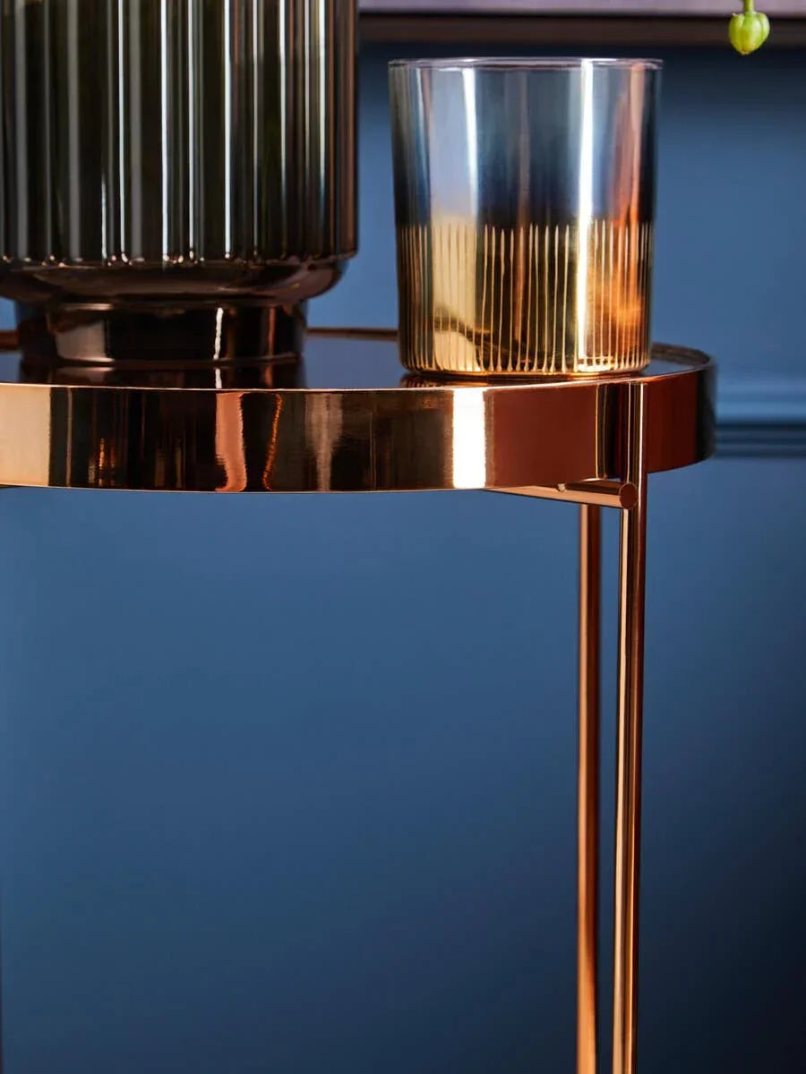 Round gold metal side table with glass candle holder and vase against blue wall