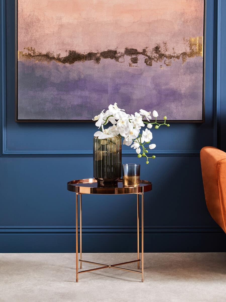 Round copper side table with glass vase, white flowers, candle, blue wall and abstract art