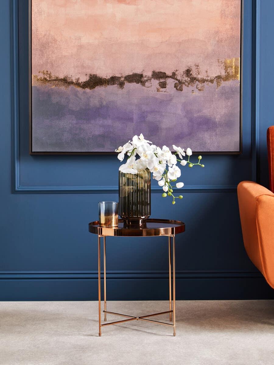 Round copper side table with glass vase of white orchids and tumbler against blue wall
