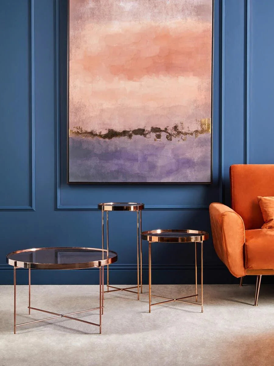 Set of three copper accent tables with glass tops beside orange armchair against blue wall