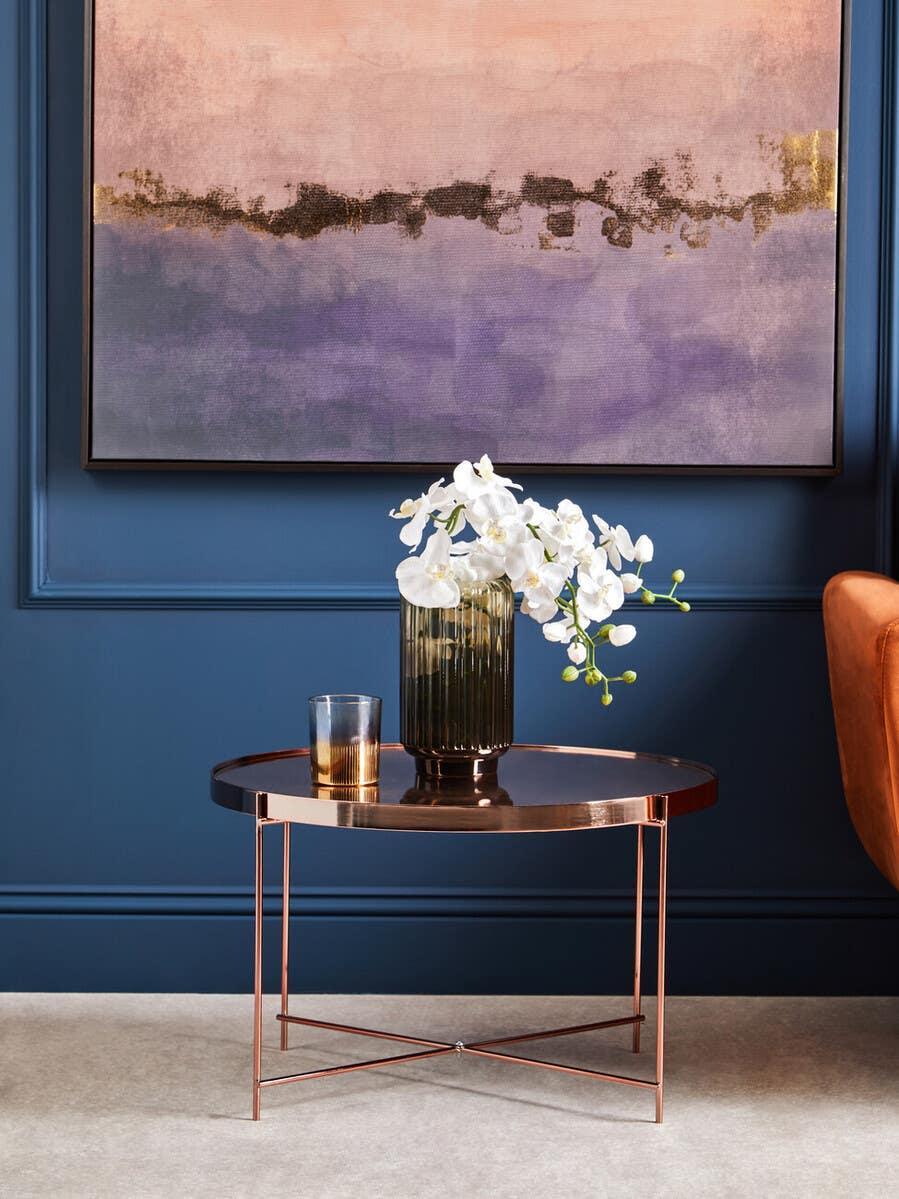 Round copper metal coffee table with glass vase and flowers against navy wall