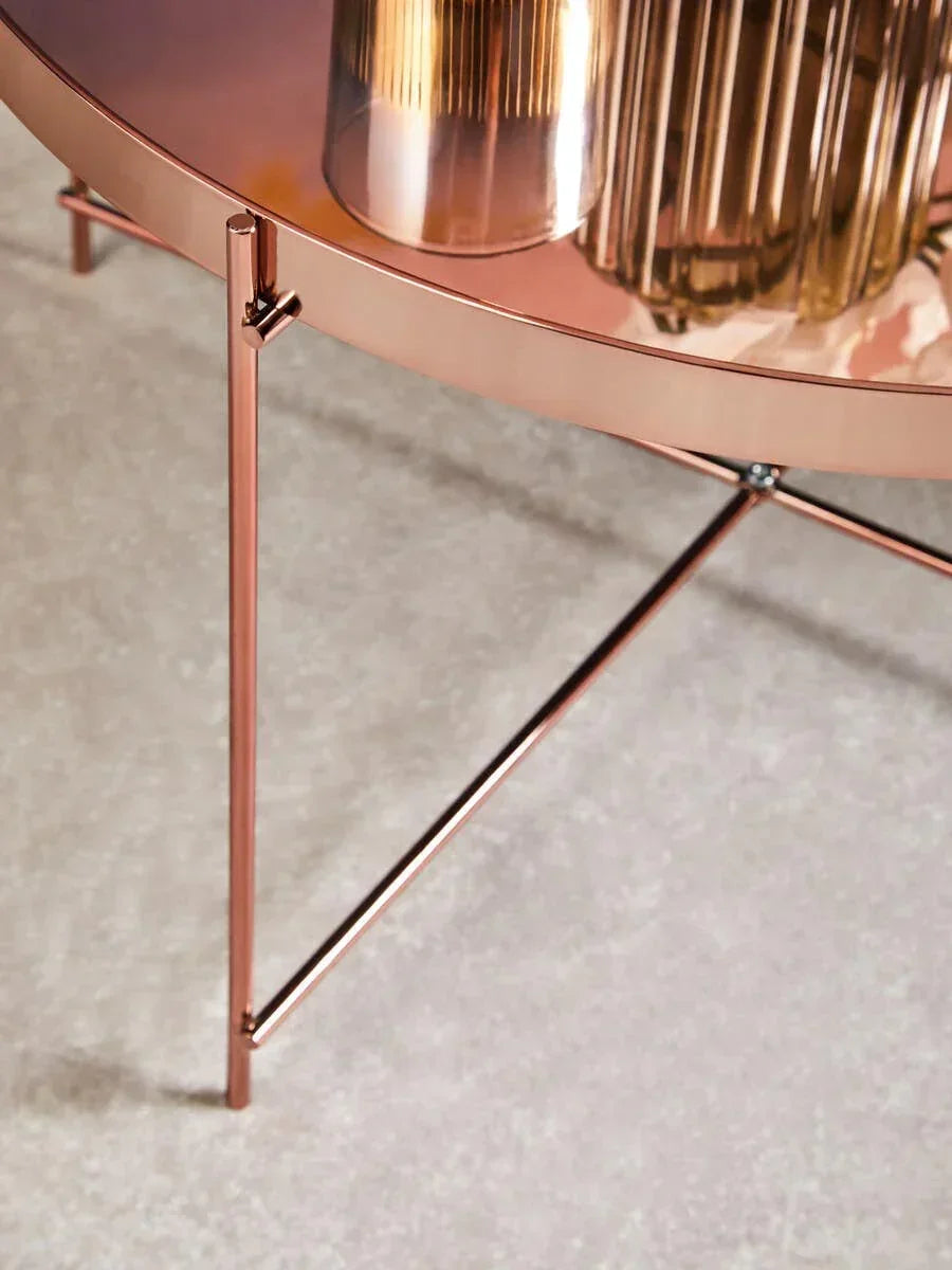 Round copper side table with metal legs and reflective glass top on a pale carpet