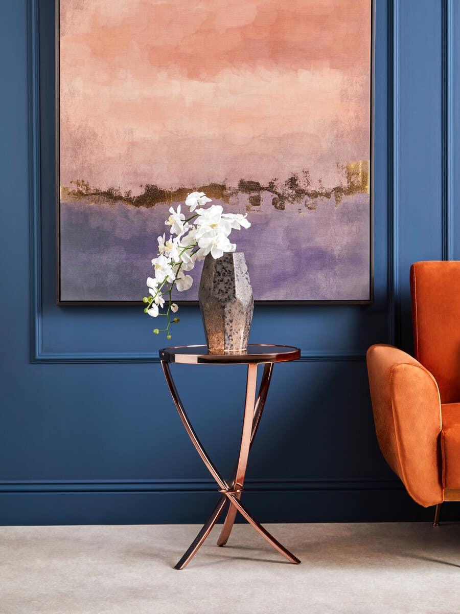 Copper metal side table with flower vase beside orange armchair against blue wall