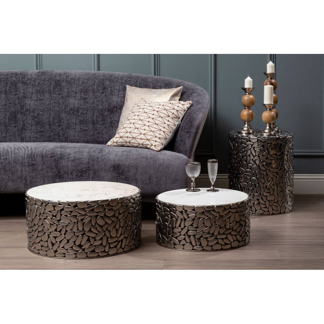 Grey velvet sofa with a curved backrest and plush cushions, accompanied by two round coffee tables with textured bases and white tops, set against a dark blue wall. A decorative pillow rests on the sofa, and a tall decorative piece with wooden accents stands beside the tables