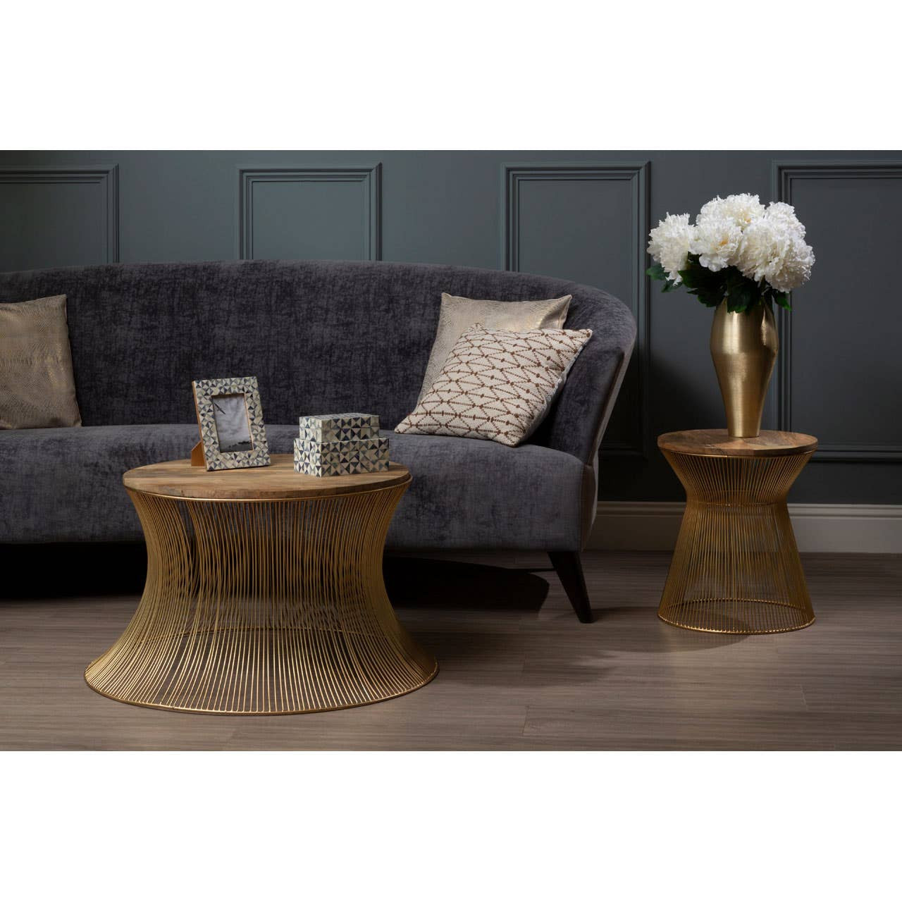 Grey velvet three-seat sofa with a textured fabric, accompanied by two woven side tables. One table holds a vase with white flowers, while the other has a picture frame. The background features a dark paneled wall, enhancing the modern aesthetic of the living space